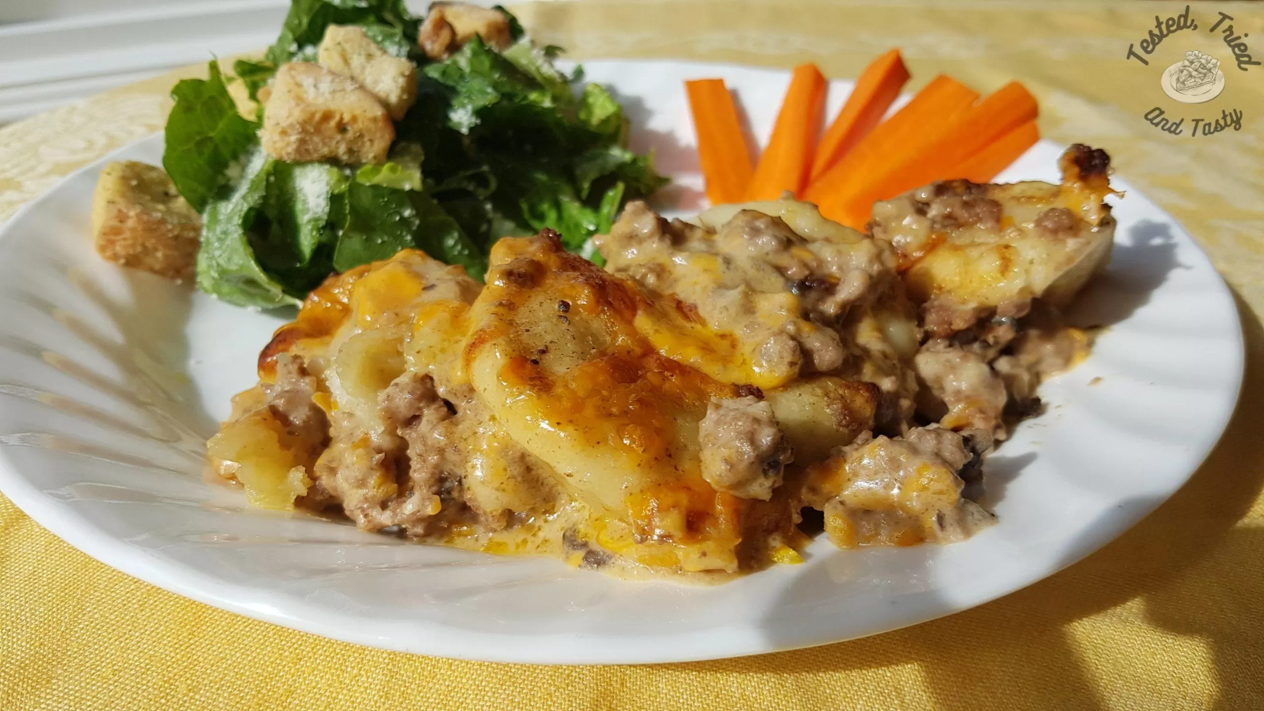 Delicious Perogy casserole with salad and carrot sticks on a white plate on a yellow tablecloth.